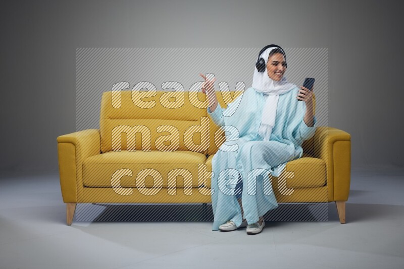 A Saudi woman wearing a light blue Abaya and a white head scarf sitting on a yellow sofa and holding an iPad while wearing headphone eye level on a grey background