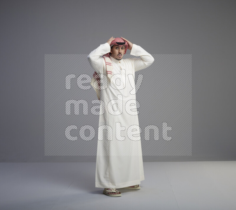 A Saudi man standing wearing thob and red shomag interacting with the camera on gray background
