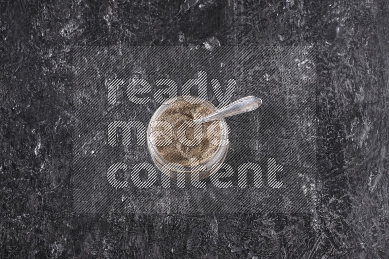 A glass jar full of black pepper powder and a metal spoon on a textured black flooring