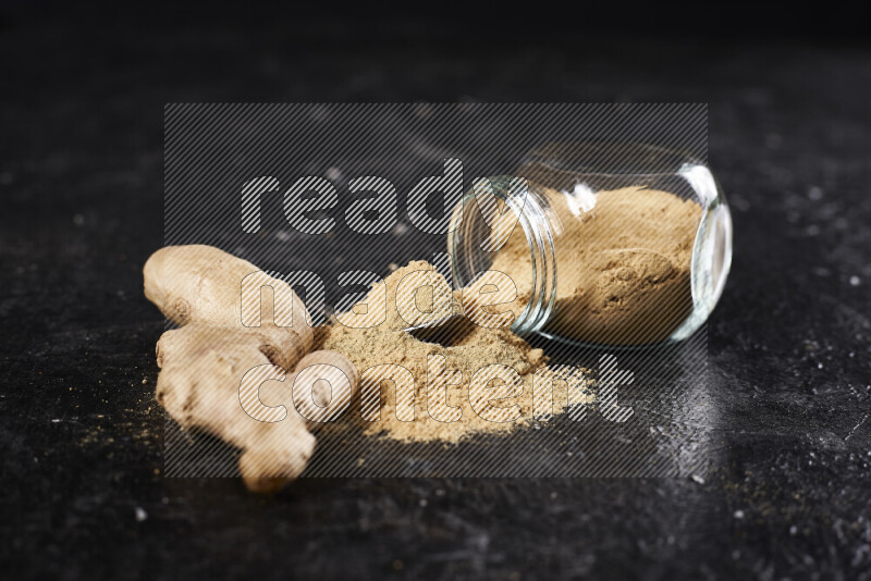 A glass jar full of ground ginger powder flipped with some spilling powder on black background
