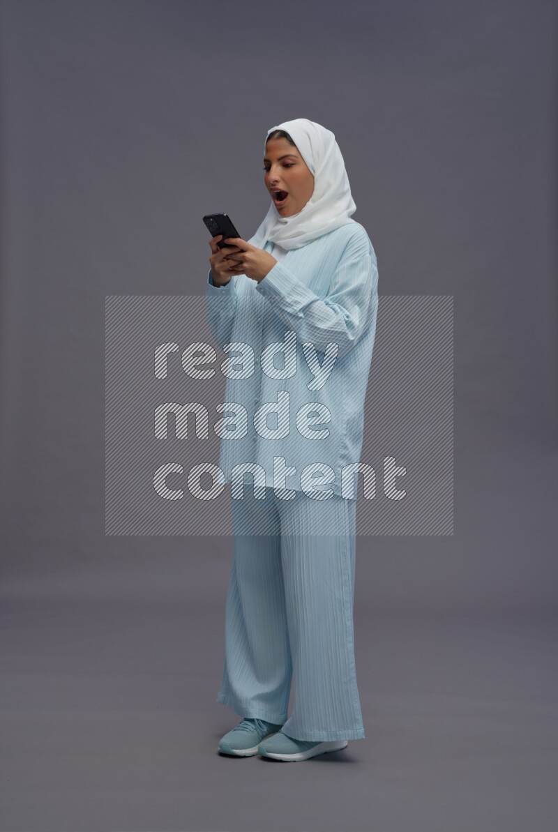 Saudi woman wearing hijab clothes standing texting on phone on gray background