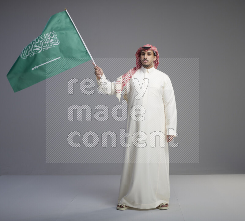 A Saudi man standing wearing thob and red shomag raising big Saudi flag on gray background