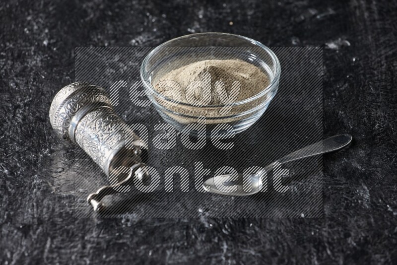 A glass bowl full of white pepper powder with pepper beads, a metal grinder and a metal spoon on textured black flooring
