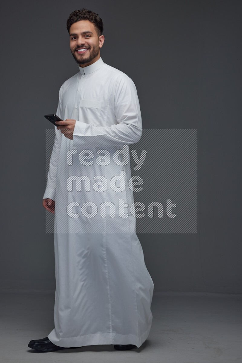 A Saudi man wearing Thobe standing and using his phone eye level on a gray background