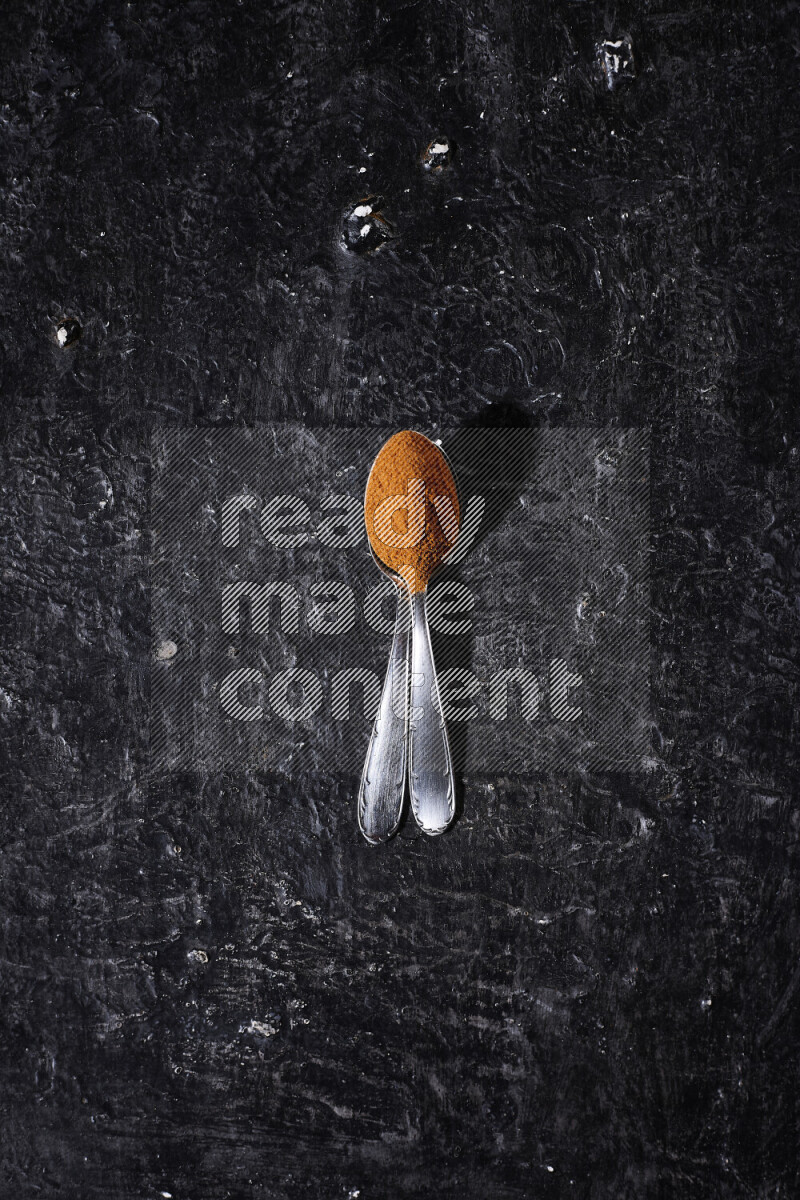 Two metal spoons full of ground paprika powder on black background