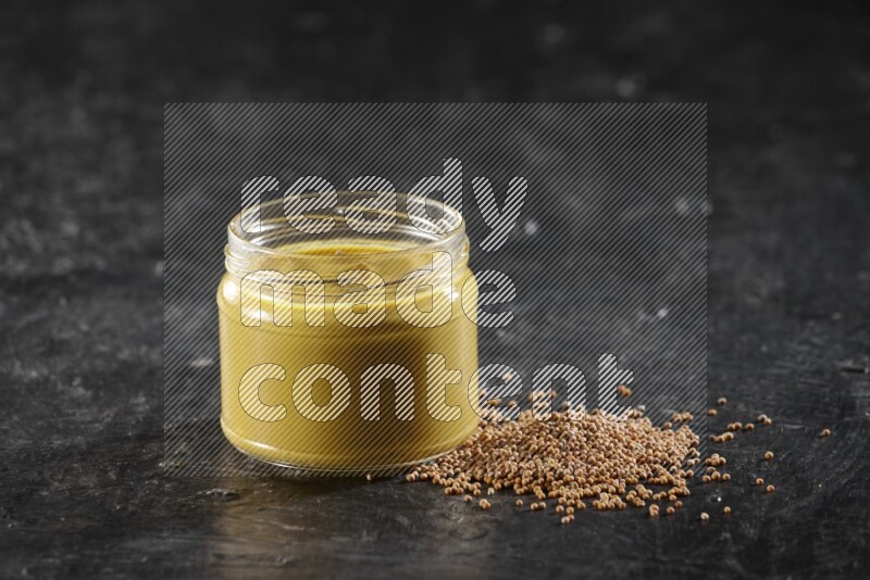 A glass jar full of mustard paste and mustard seeds spread next to it on a textured black flooring