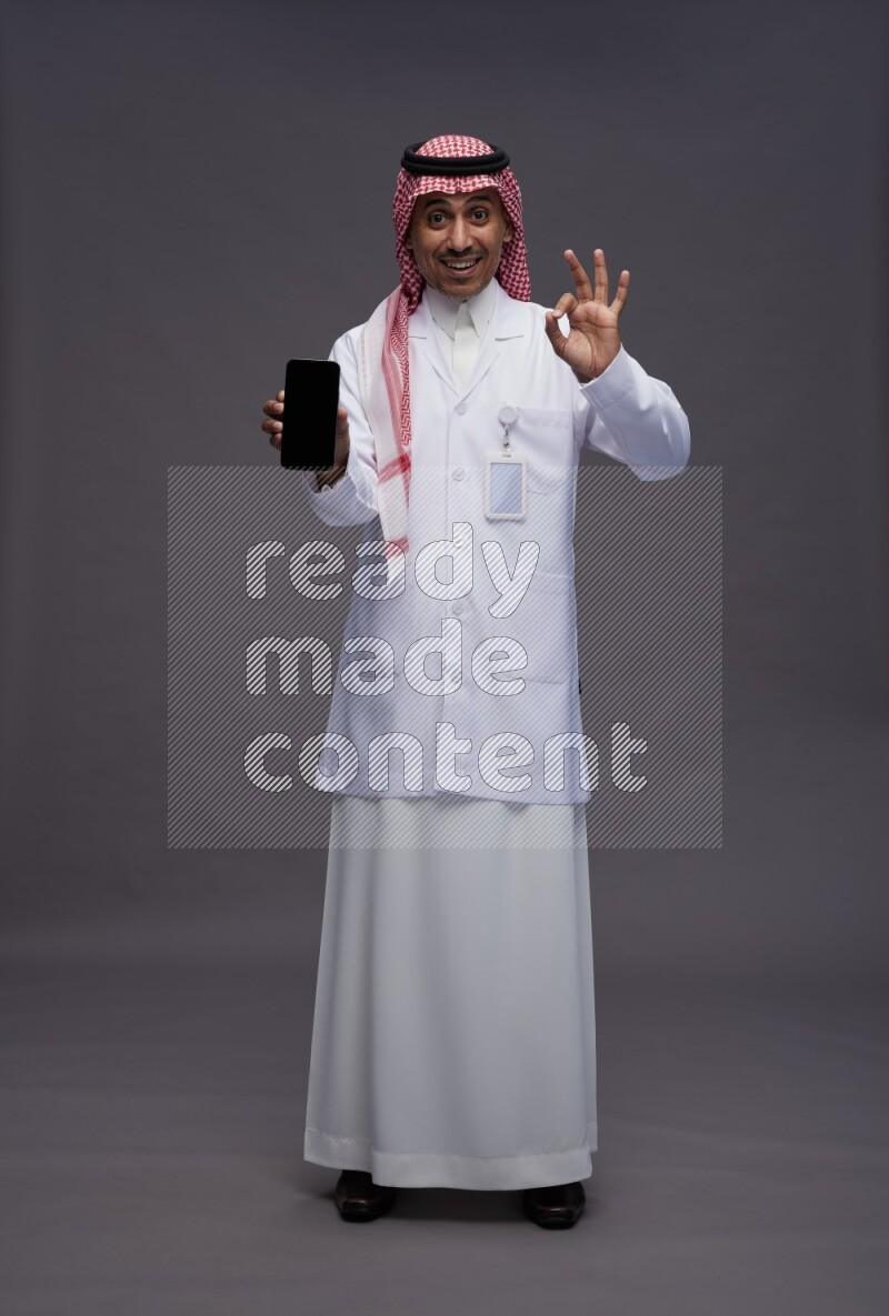 Saudi man wearing thob with lab coat and shomag with pocket employee badge standing showing phone to camera on gray background