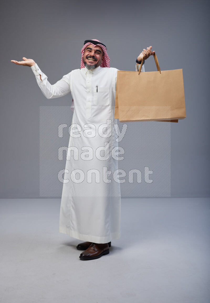 Saudi man Wearing Thob and red Shomag standing holding shopping bag on Gray background