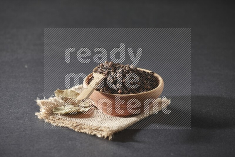 A wooden bowl, a wooden spoon full of cloves, and bay leaves (laurel) on a piece of burlap on a black flooring