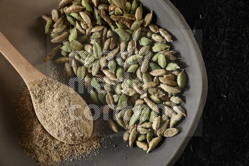 A plate filled with cardamom seeds and a wooden spoon full of cardamom powder on a textured black flooring