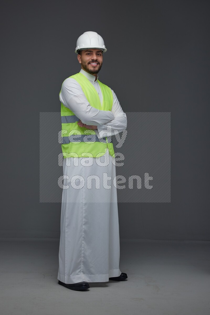 A Saudi man wearing Thobe with a yellow safety vest and white helmet standing and crossing his hands eye level on a gray background