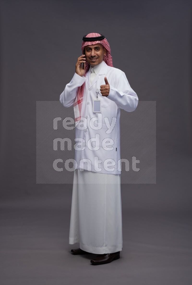 Saudi man wearing thob with lab coat and shomag with pocket employee badge standing talking on phone on gray background