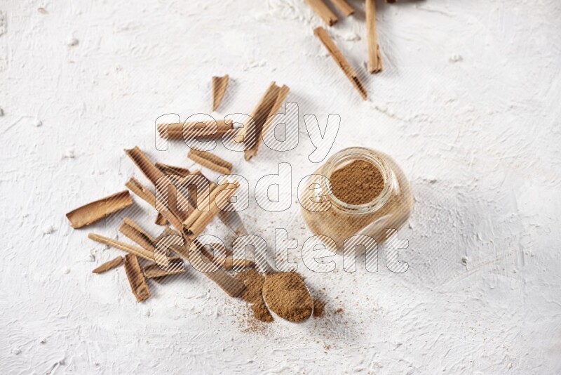 Herbal glass jar full cinnamon powder and a metal spoon surrounded by cinnamon sticks on a white background