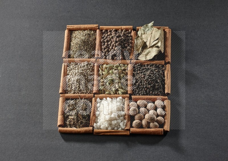 9 squares of cinnamon sticks full of cardamom in the middle surrounded by nutmeg, black pepper, bay laurel leaves, allspice, cumin, white pepper, dried basil and salt on black flooring