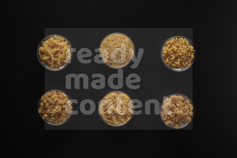 Different pasta types in 6 glass bowls on black background