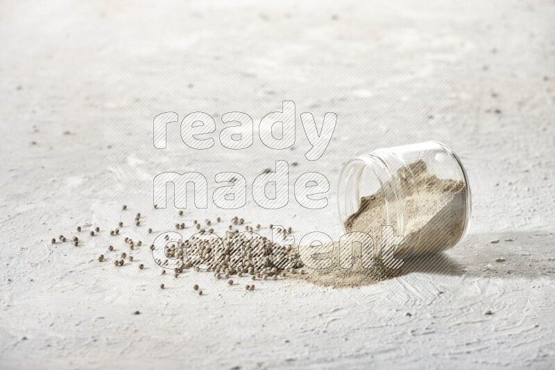 A flipped glass jar full of white pepper powder with spilled powder and white pepper beads on textured white flooring