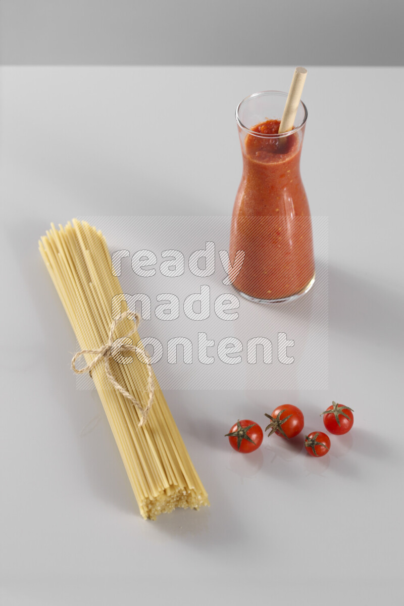 Raw pasta with tomatoe pasta with different ingredients such as cherry tomatoes, basil, garlic, bay laurel, cardamom, white pepper, black pepper, red chilis and wheat stalks on light grey background