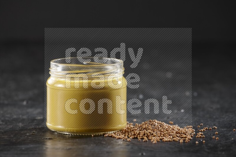 A glass jar full of mustard paste and mustard seeds spread next to it on a textured black flooring