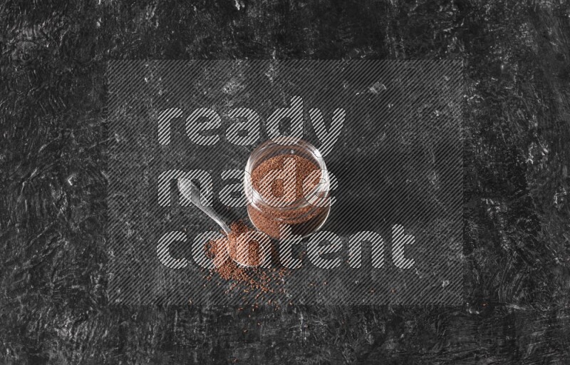 A glass jar full of garden cress seeds with a metal spoon full of the seeds on a textured black flooring