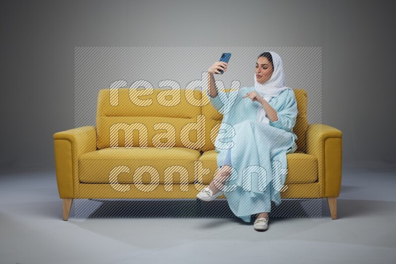A Saudi woman wearing a light blue Abaya and white head scarf sitting on a yellow sofa and taking selfies with her phone eye level on a grey background