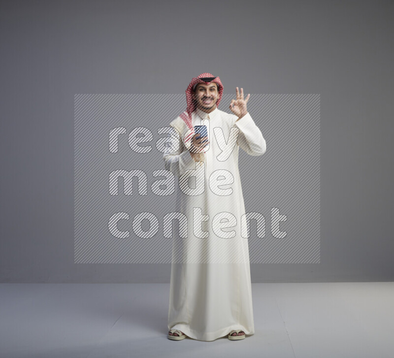 A  Saudi man standing wearing thob and red shomag texting on phone on gray background