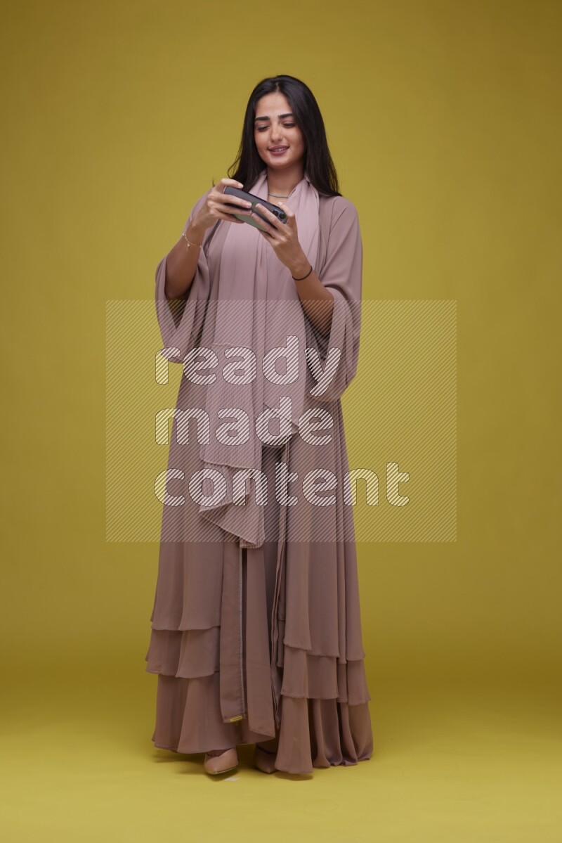 A woman Playing a Game on her smartphone on a Yellow Background wearing Brown Abaya