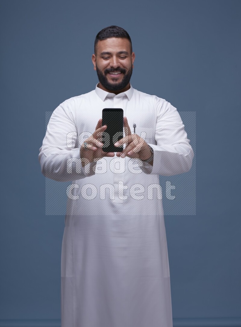 A man Showing his phone in Blue Background wearing Saudi Thob