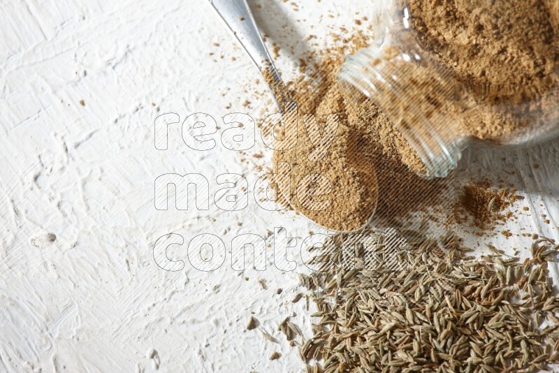 A glass spice jar and metal spoon full of cumin powder and the jar flipped and powder spilled out with cumin seeds on textured white flooring