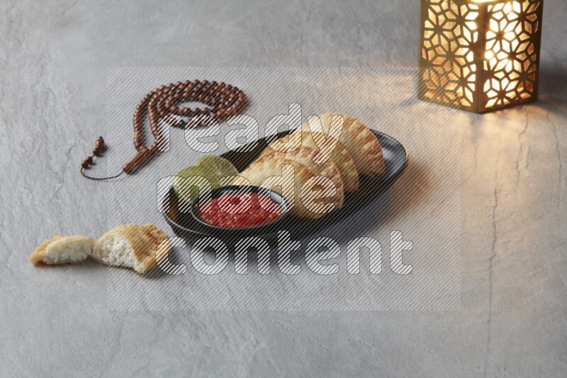 Four fried sambosas in an oval shaped black plate, beside a cut cheese sambosa, a brown misbaha and a golden lantern on a gray background