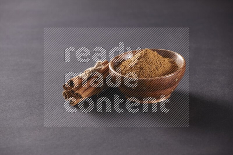 Cinnamon sticks stacked and bounded beside a wooden bowl full of cinnamon powder and a wooden spoon full of powder on black background