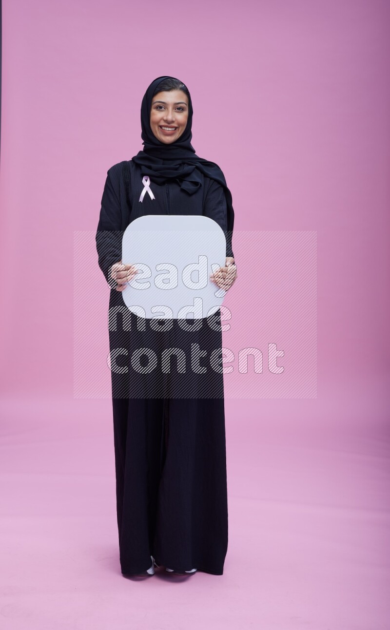 Saudi woman wearing pink ribbon on Abaya standing holding social media signs on pink background