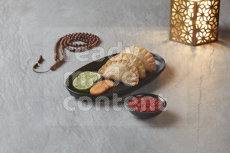 Three fried sambosas in an oval shaped black plate and a red sauce in a black round ramekin with a brown misbaha and a golden lantern on the side on a gray background