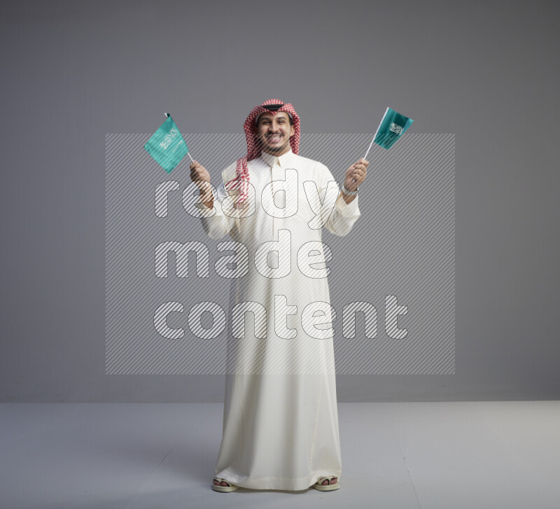 A Saudi man standing wearing thob and red shomag raising small Saudi flag on gray background