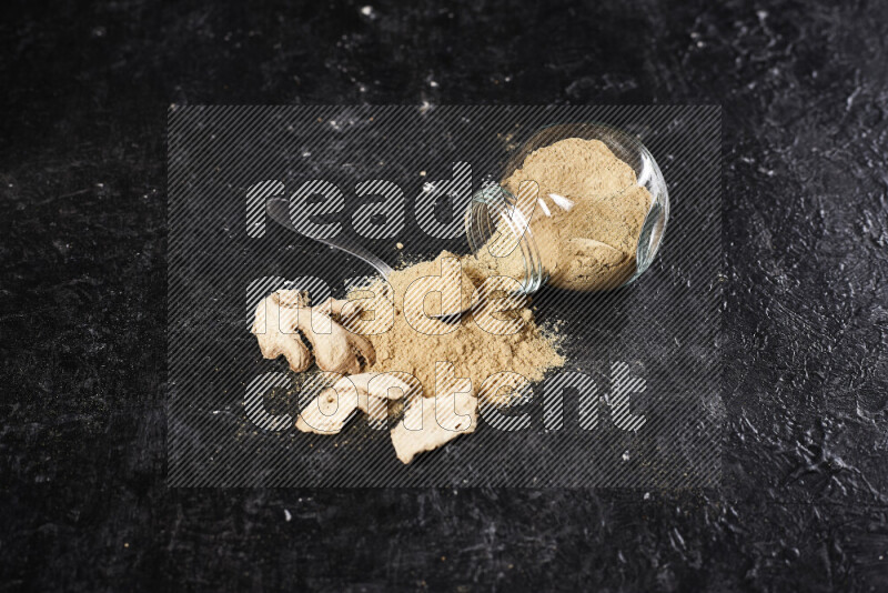 A glass jar full of ground ginger powder flipped with some spilling powder on black background