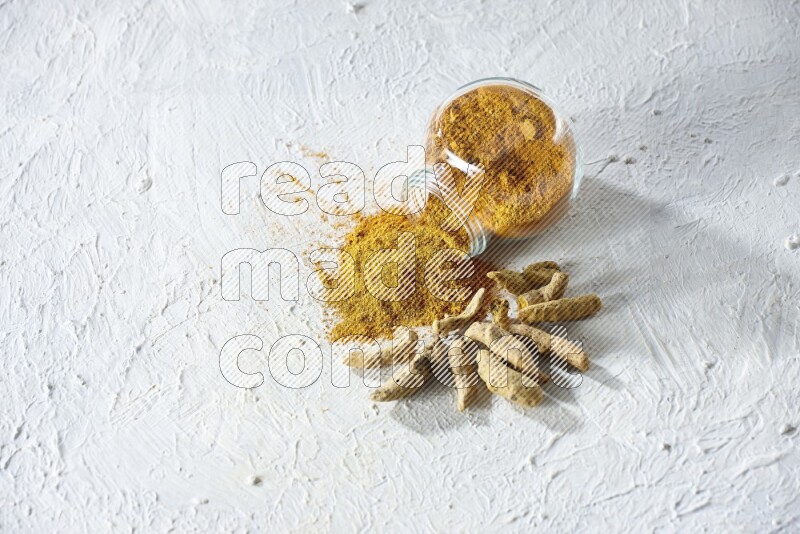 A flipped glass spice jar full of turmeric powder and powder spilled out of it with dried whole fingers on textured white flooring