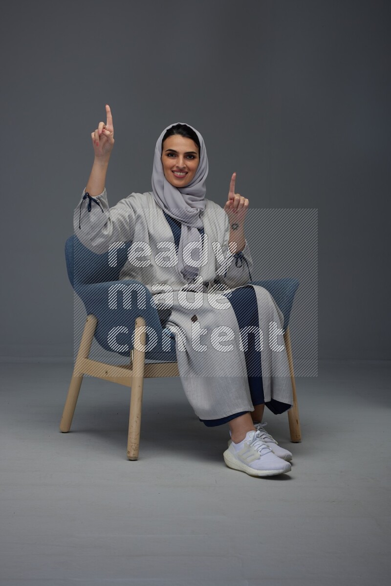 A Saudi woman wearing a light gray Abaya and head scarf sitting on a dark grey chair while making multi hand gestures eye level on a grey background