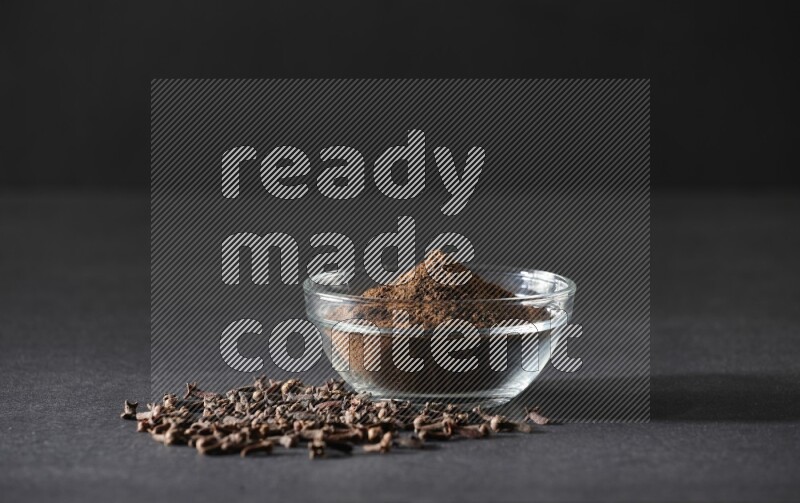 A glass bowl full of cloves powder with cloves grains spread on black flooring