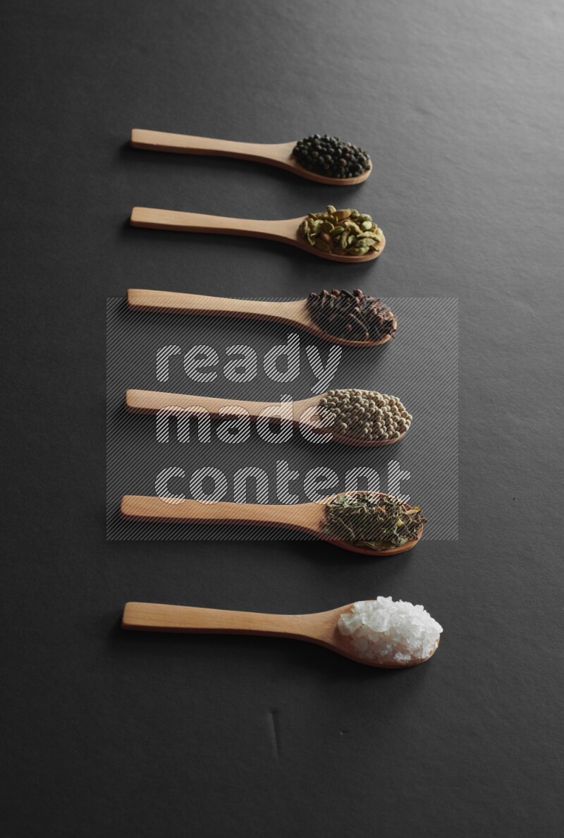 wooden spoons filled with white peppers, cloves, cardamom, salt, black peppers and basil on black flooring