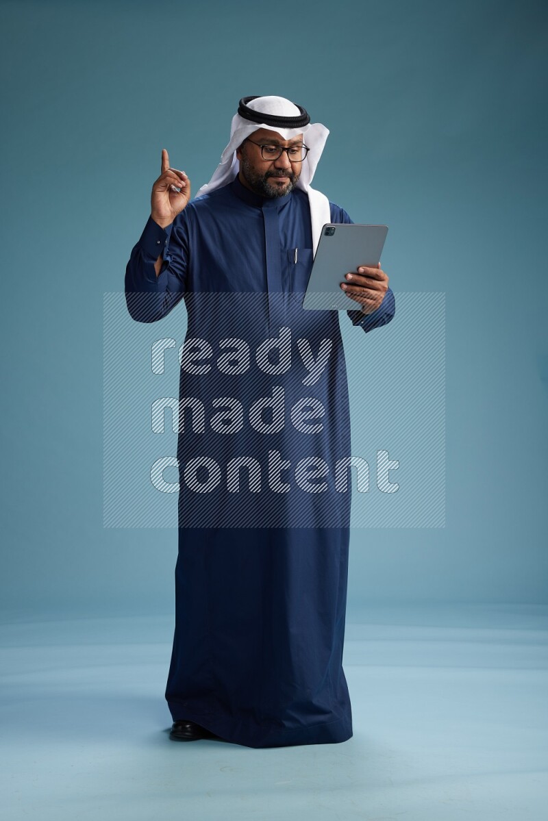 Saudi Man with shimag Standing holding ATM card while working on tablet on blue background
