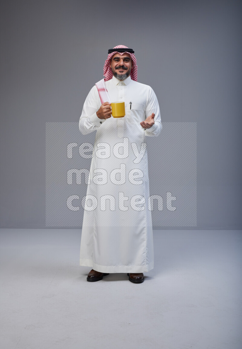 Saudi man Wearing Thob and red Shomag standing holding mug on Gray background