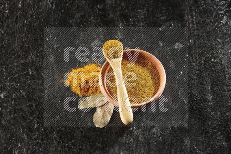 A wooden bowl and a wooden spoon full of turmeric powder on textured black flooring