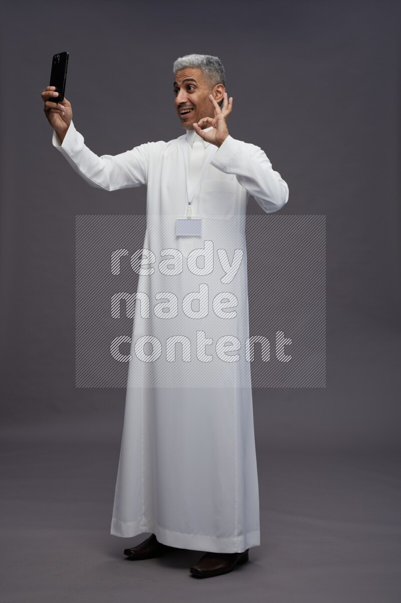 Saudi man wearing thob with neck strap employee badge standing taking selife on gray background