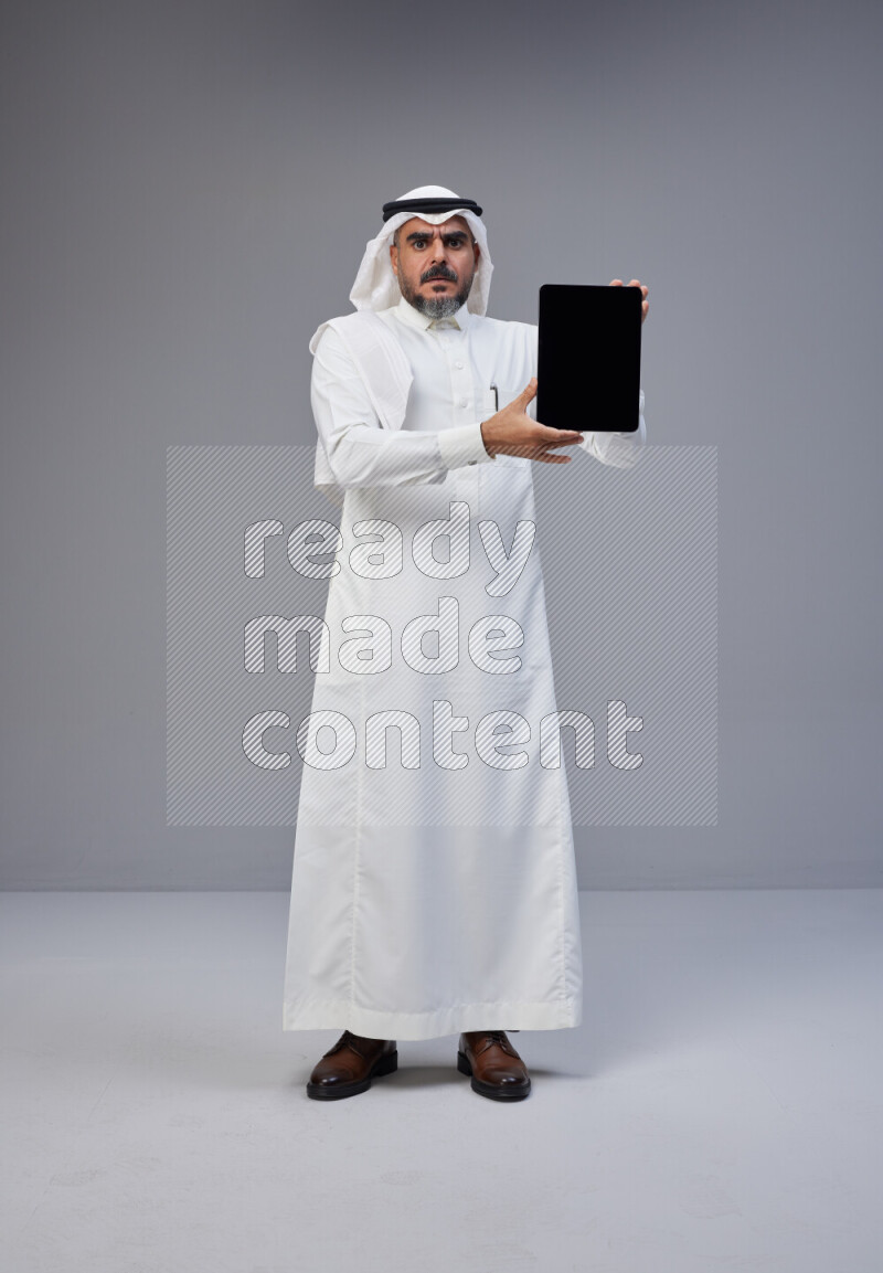 Saudi man Wearing Thob and white Shomag standing showing tablet to camera on Gray background