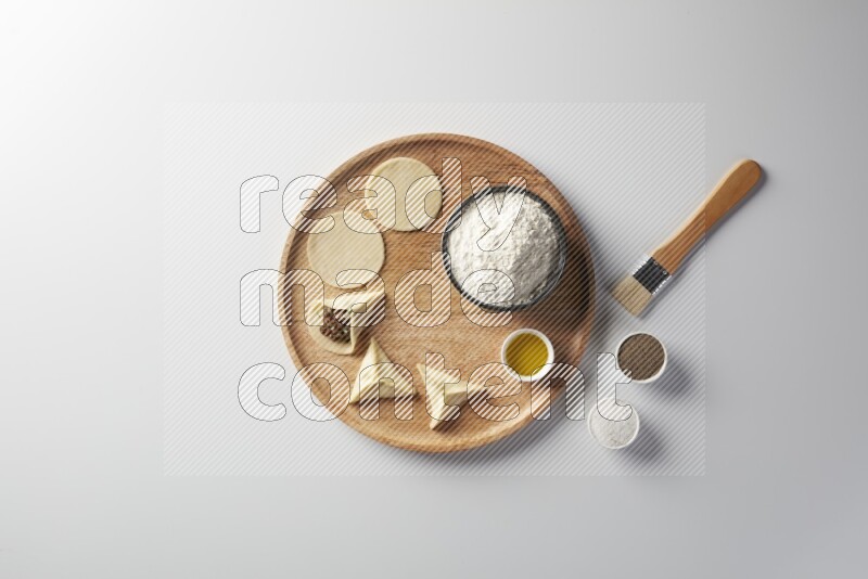 two closed sambosas and one open sambosa filled with meat while flour, salt, black pepper and oil with oil brush aside in a wooden dish on a white background