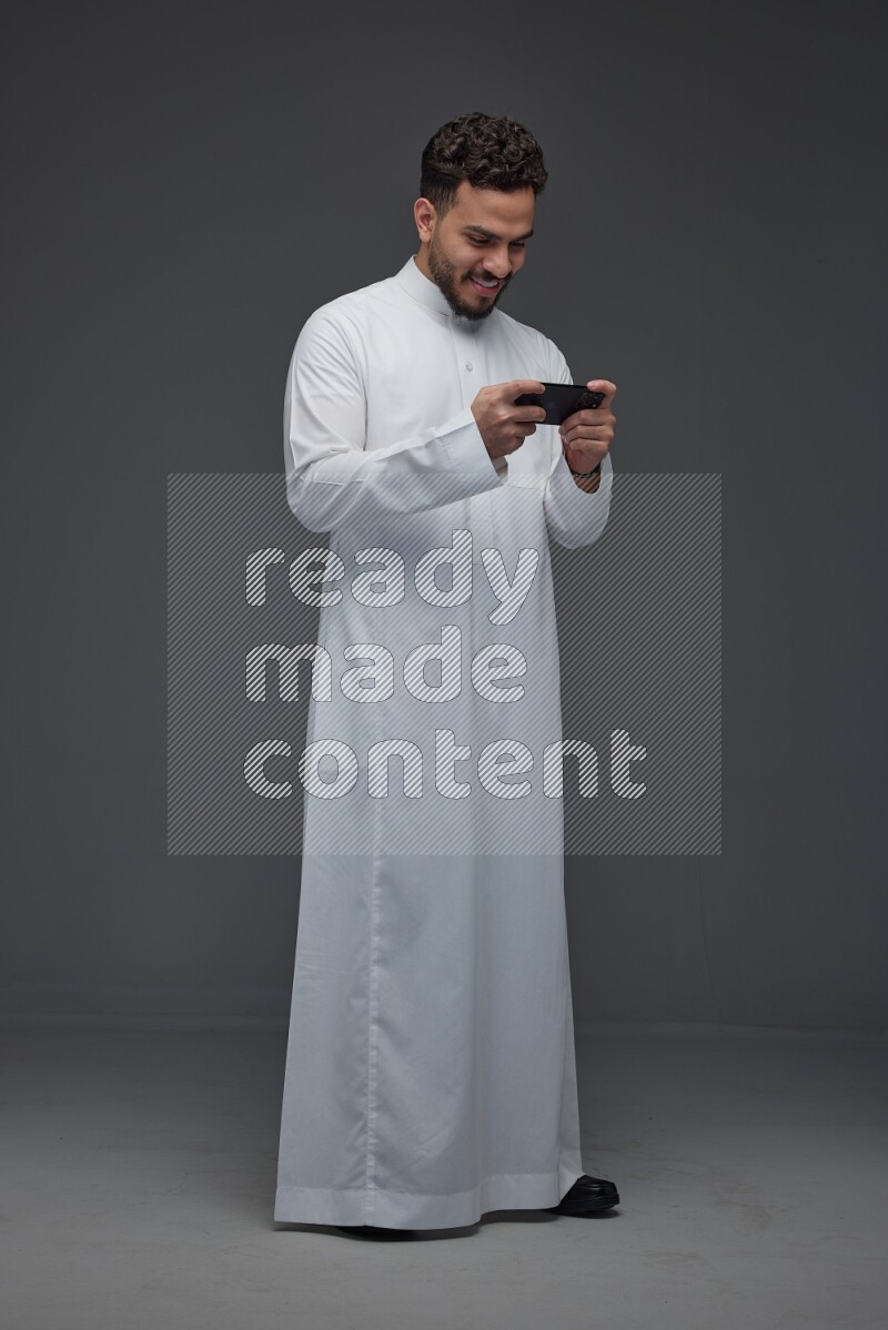 A Saudi man wearing Thobe and playing using his phone horizontally playing video games while standing and making different poses eye level on a gray background