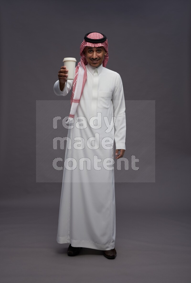 Saudi man wearing thob and shomag standing holding paper cup on gray background