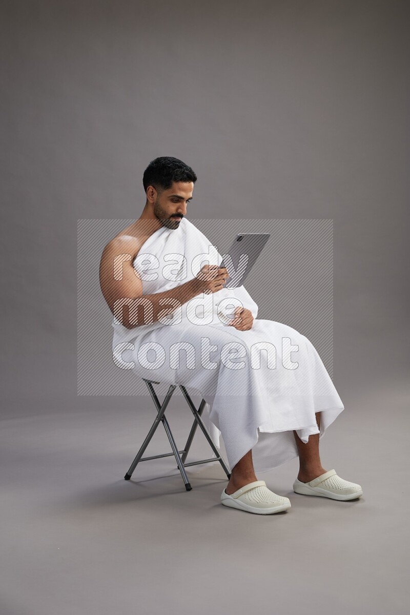 A man wearing Ehram sitting on chair holding a tablet on gray background