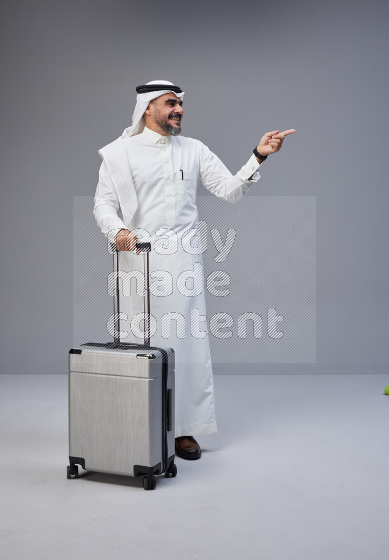 Saudi man wearing Thob and white Shomag standing holding Travel bag on Gray background