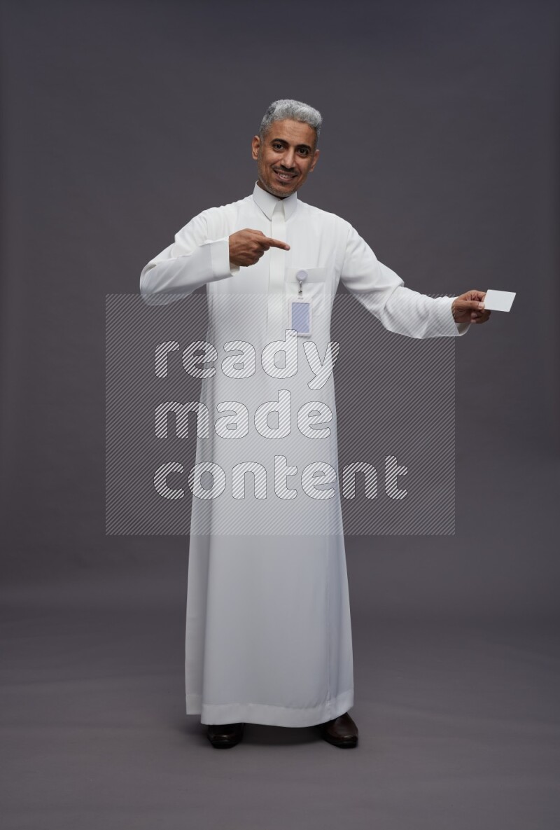 Saudi man wearing thob with pocket employee badge standing holding ATM card on gray background
