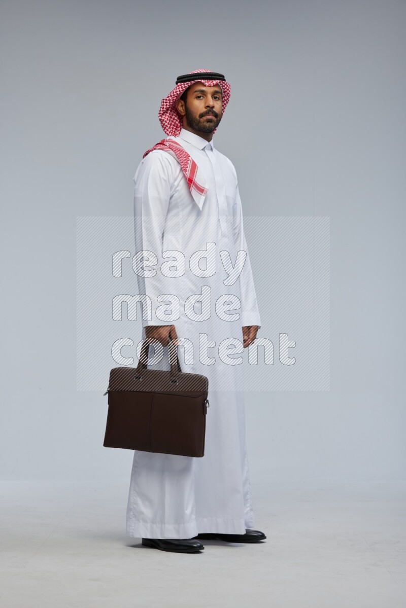 Saudi man Wearing Thob and shomag standing holding bag on Gray background